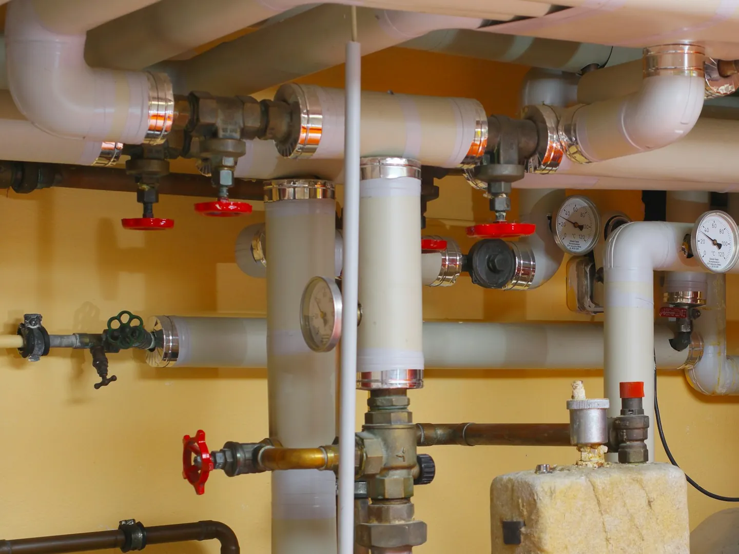 A plumbed mechanical room: insulated hot-water lines, red shut-off valves and temperature gauges on a warm-painted wall